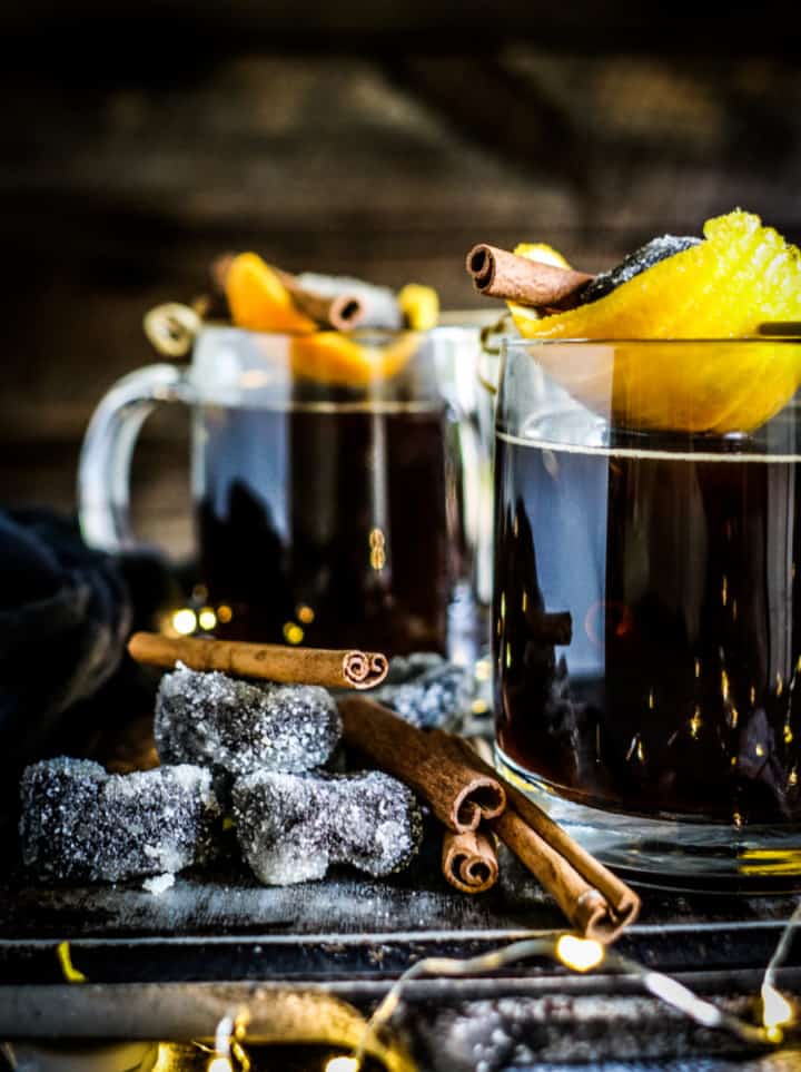 Two glass mugs containing a dark drink with lemon peel and cinnamon sticks on top.