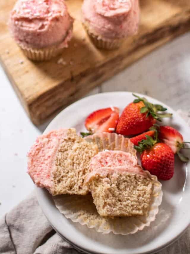 one cupcake cut in half on a white plate with strawberries garnishing the side - two whole cupcakes in the background.