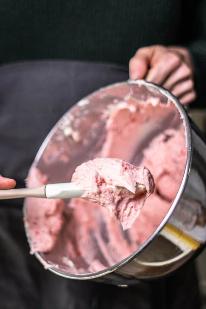 a silver mixing stand bowl with pink strawberry frosting inside being scooped out with a spatula 