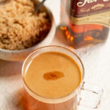 A mug full of hot buttered rum next to a bottle of rum, & a bowl of brown sugar. All on. a white back ground
