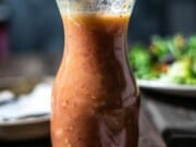 Glass bottle filled with tomato vinaigrette in front of a salad on a wooden table.
