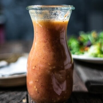 Glass bottle filled with tomato vinaigrette in front of a salad on a wooden table.