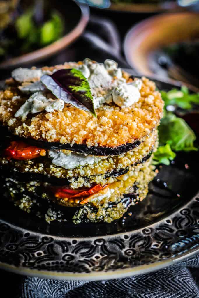 Stack of eggplant napoleon on a dark grey plate with a bed of greens underneath it