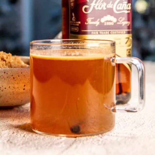 Clear mug full of hot buttered rum with a bottle of dark rum in the background