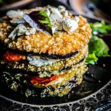 Stack of eggplant napoleon on a dark grey plate with a bed of greens underneath it