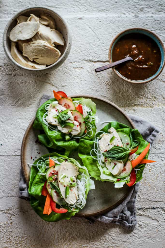 Close-up shot of 3  plated chicken lettuce wraps with a side of peanut sauce & a bowl of sliced chicken  all on a white wooden table