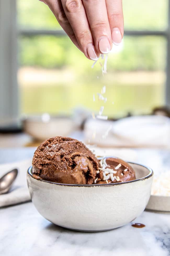 A scoop of ice cream in a bowl with a hand sprinkling coconut on top