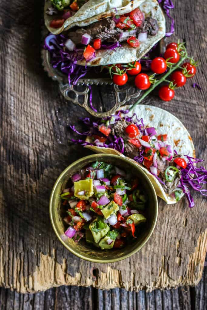 steak tacos on a wooden background with a bowl of avocado salsa & fresh tomatoes
