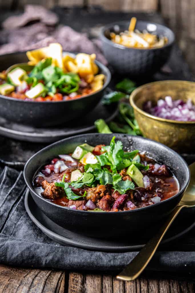 two bowls of homemade chili surrounded by bowls of chili toppings